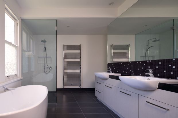 A bright contemporary bathroom with a freestanding white bathtub, twin white vessel basins on a long pale vanity, large wall mirrors, a glass-enclosed shower, heated towel rails, dark tiled flooring, and a glossy dark mosaic tile backsplash. Natural light enters through the large window on the left.