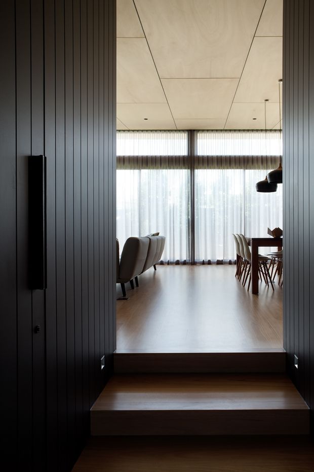 A refined modern interior viewed from a darker lowered entry or hallway, looking up two broad timber steps into an open-plan living and dining area. Dark vertical wall panelling frames the opening, while a pale timber-panelled ceiling, warm timber flooring, floor-to-ceiling sheer curtains, sculptural pendant lighting, a sofa, dining table and upholstered dining chairs create a calm, softly lit composition.