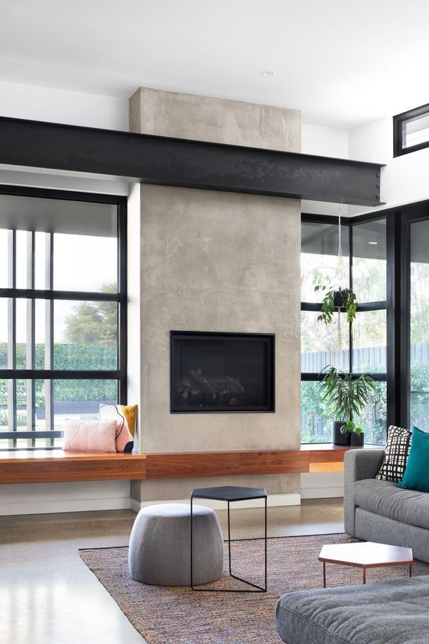 Bright contemporary living room with a central textured concrete or plaster fireplace wall, inset black fireplace, and a projecting dark steel structural beam. Floor-to-ceiling black-framed glazing surrounds a built-in timber window seat, while a grey sofa, ottoman, small black side table, patterned rug, and teal cushion furnish the room. Indoor plants and a garden are visible through the windows.