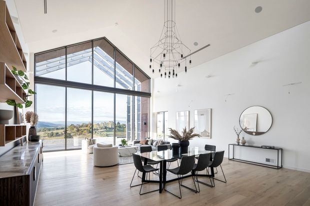 A bright, spacious open-plan living and dining area with a dramatic double-height glazed wall overlooking a landscaped suburban setting. The room features pale timber flooring, white walls, extensive black-framed glazing, built-in timber shelving and cabinetry, a contemporary black dining table with matching chairs, a minimalist sideboard, and a sculptural suspended chandelier. A lounge area is visible beyond the dining zone.