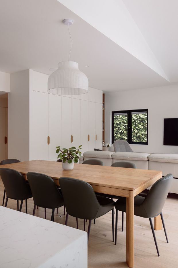 A bright, open-plan dining and living area with a large light timber dining table surrounded by dark upholstered chairs. White built-in cabinetry with slim timber handles lines the rear wall, while a cream sectional sofa, black-framed window, wall-mounted television, and oversized ribbed pendant light complete the space. A white stone kitchen island is visible in the foreground. The room has a clean, softly minimalist contemporary appearance.