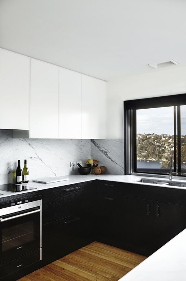 A bright, compact contemporary kitchen with matte black lower cabinetry, handleless white overhead cabinets, a white veined stone splashback and benchtop, stainless-steel appliances, and warm timber flooring. A large black-framed window overlooks a leafy hillside and water beyond.