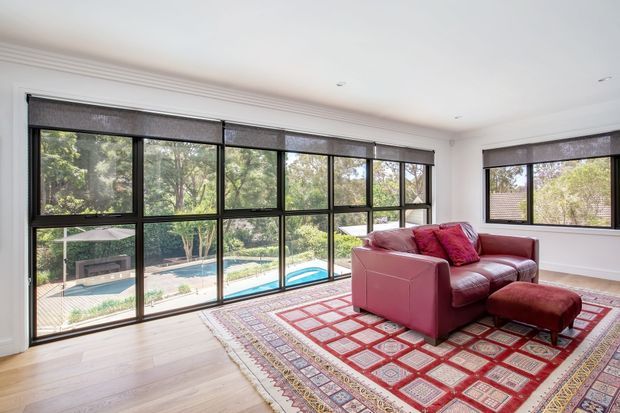 Bright contemporary living room with full-height black-framed glazing overlooking a landscaped garden and swimming pool. The room has pale timber flooring, white walls with decorative cornice detailing, recessed ceiling lights, roller blinds, and a burgundy leather sofa with matching ottoman positioned on a large patterned red rug.