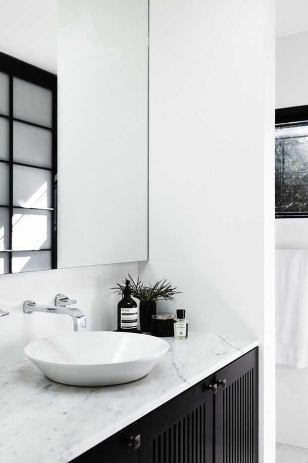 A bright contemporary bathroom vanity with a white vessel basin set into a white marble-look benchtop. The vanity has dark, vertically fluted cabinetry, a wall-mounted chrome tap, a large mirrored cabinet, and black-framed windows. Minimal toiletries and a small plant sit on the counter.