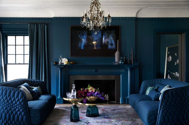 A richly styled, symmetrical living room in deep navy blue, featuring two quilted blue sofas facing a classical fireplace, ornate crystal chandelier, dark wall panelling, framed artwork, patterned rug, and small sculptural coffee tables. Tall curtained windows provide natural light, creating a dramatic and luxurious atmosphere.