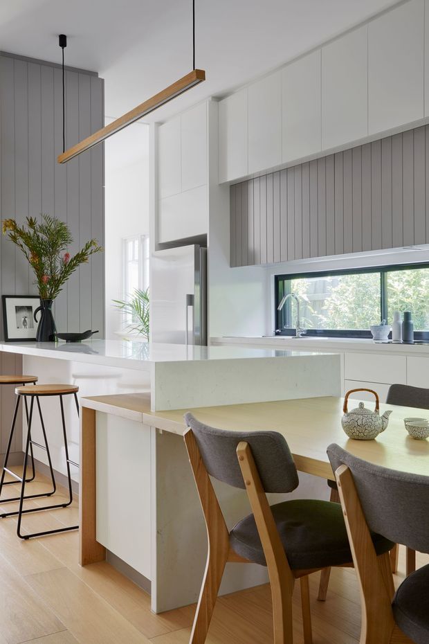 Bright contemporary kitchen and dining area with a large white stone island, integrated white cabinetry, grey vertical-panel feature walls and splashback, timber flooring, and a linear timber pendant light. A dining table with upholstered timber-framed chairs occupies the foreground, while stools sit at the island. The room has abundant natural light through a large kitchen window and glazed opening.