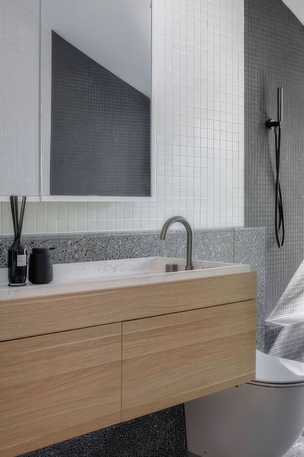 Contemporary bathroom vanity area featuring a floating light timber double-drawer vanity, white integrated basin, brushed metal curved faucet, grey terrazzo-style benchtop and wall band, and small-format white and charcoal grid tiles. A dark shower fixture and freestanding bathtub are visible on the right, with a framed geometric mirror above the vanity.