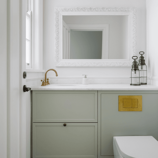A refined compact bathroom vanity with pale sage-green built-in cabinetry, a white stone countertop, brass tapware, ornate white-framed mirror, wall panelling, and a partially visible toilet. The space has a soft, classic decorative character with restrained contemporary detailing.