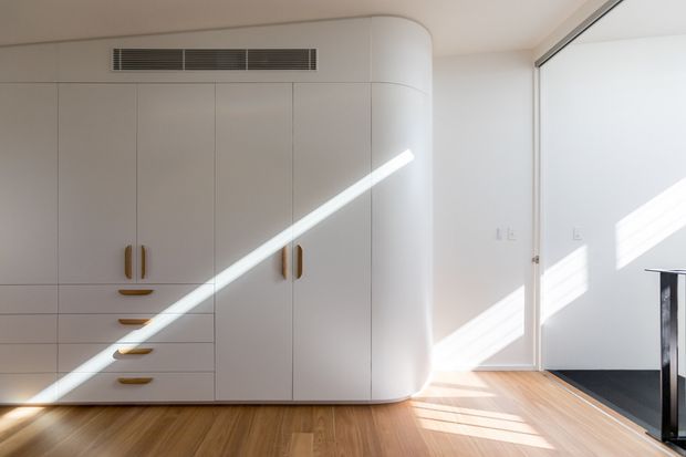 Bright minimalist interior with a large wall of custom curved cabinetry in a white finish, integrated drawers and doors with warm brass-toned handles, and a warm timber floor. A narrow doorway and strong sunlight enter from the right, creating diagonal light across the cabinetry and floor.