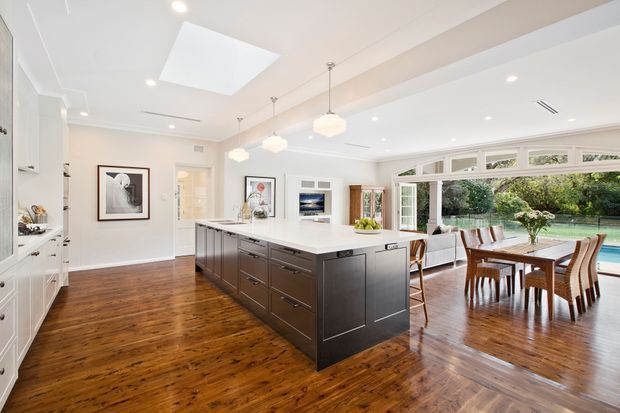 Bright open-plan kitchen and dining area with a large dark-stained island, white cabinetry and benchtops, warm timber flooring, pendant lights, skylight, and extensive glazed doors opening to a leafy outdoor area. A dining table and part of a living area are visible beyond the kitchen.