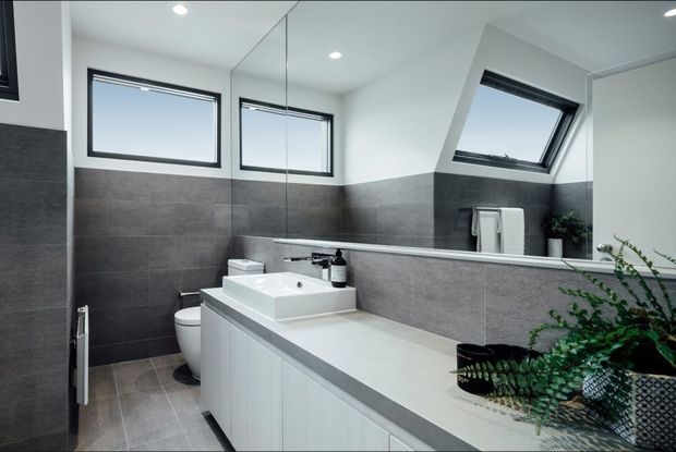 Contemporary bathroom with a long white vanity and integrated basin, grey tiled walls and floor, frameless shower glazing, wall-mounted towel rail, skylight-style windows, and a large mirrored wall reflecting the vanity and toilet.