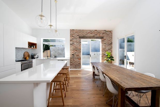 Bright open-plan kitchen and dining area with white contemporary cabinetry, a large white island, patterned tile splashback, timber flooring, exposed brick feature wall, pendant lights, and a long rustic timber dining table. Glazed doors and windows provide abundant natural light and connect to an outdoor area.