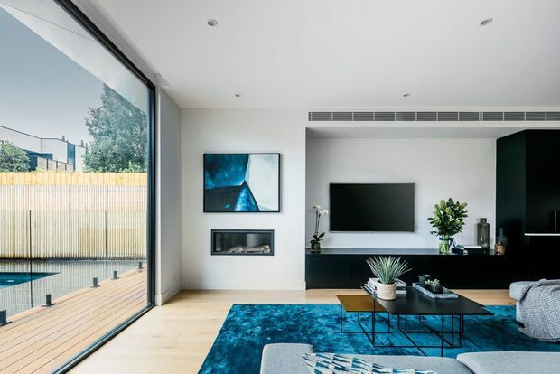 Bright contemporary living room with pale timber flooring, white walls and ceiling, a large corner glazed opening to a timber deck and pool, and a minimalist black built-in media wall. A teal-blue rug, abstract artwork, low black coffee tables, indoor plants and a light sectional sofa add colour and texture.