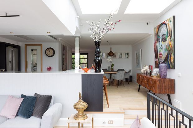 Bright open-plan residential interior viewed from a slightly raised living area, with a white sofa in the foreground, a compact dark kitchen island, dining area, timber sideboard, colourful portrait artwork, decorative objects, and large skylights. A black metal balustrade separates the living zone from the dining and kitchen areas.