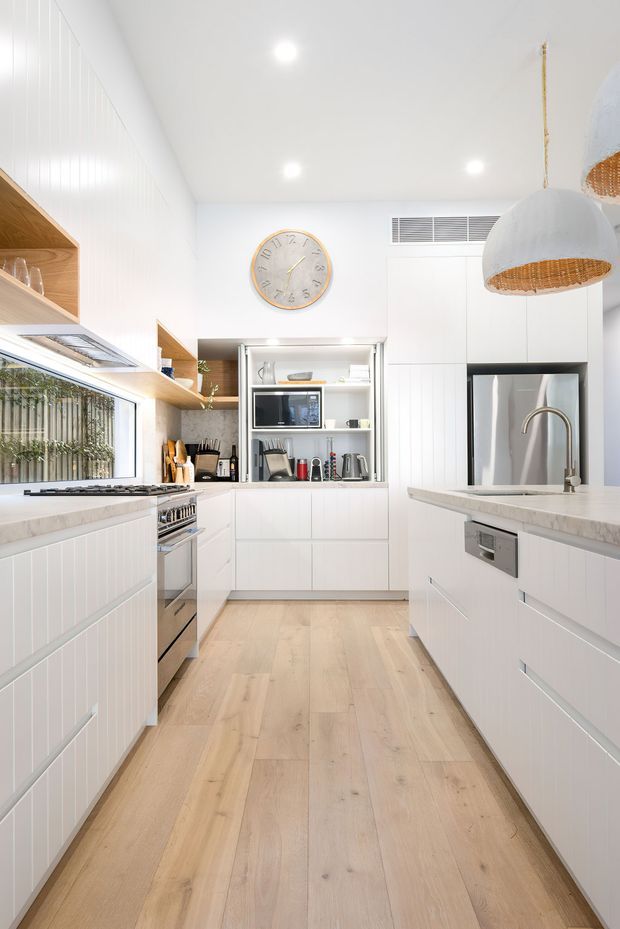 Bright, contemporary galley kitchen with white vertical-panel cabinetry, pale timber flooring, light stone benchtops, integrated stainless-steel appliances, open timber shelving, and a central white display and appliance cabinet. A large window provides natural light, while woven pendant lights and a round wall clock add warmth and texture.