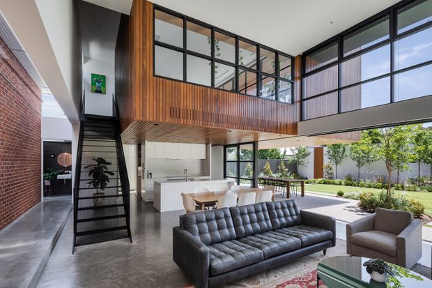 A spacious double-height contemporary living area with an open-plan kitchen and dining zone. The room features a black tufted leather sofa, upholstered armchairs, a white island kitchen, polished concrete flooring, extensive black-framed glazing, and a dramatic upper-level timber-lined volume with an internal stair and mezzanine-like overlook. A brick wall and landscaped courtyard are visible beyond the open glazing.