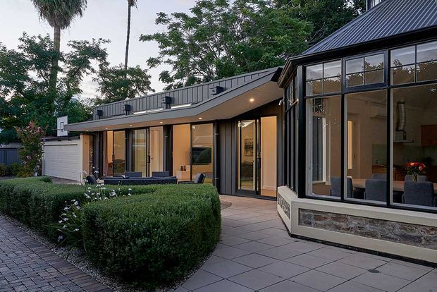 A modern single-storey residence viewed from a landscaped courtyard, with dark standing-seam metal cladding and roofing, extensive black-framed glazing, warm interior lighting, a low-pitched connecting roof, and a glazed projecting pavilion. Formal clipped hedges, tiled paving, and a small outdoor seating area frame the building.
