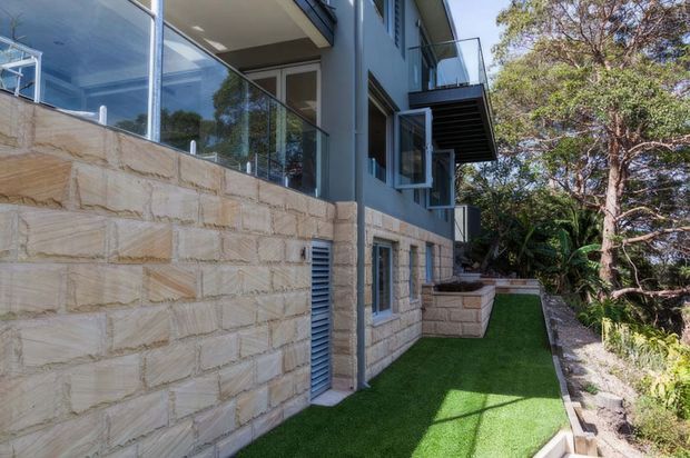Exterior side view of a contemporary multi-level residence, featuring pale textured stone blockwork at the lower level, dark blue-grey upper walls, extensive glazing with glass balustrades, projecting balconies, and a narrow landscaped side yard with artificial turf.