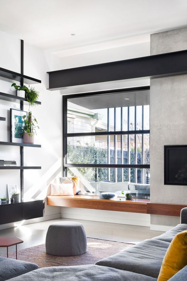 Bright contemporary living room with a large black-framed window, integrated timber window seat, built-in open shelving, a textured concrete-look feature wall with wall-mounted television, and a large grey sectional sofa. Strong sunlight creates an airy, modern interior with black structural and joinery accents.