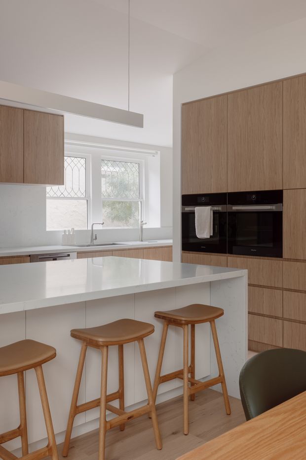 A bright, refined contemporary kitchen featuring extensive light timber cabinetry, integrated black appliances, white stone surfaces, and a large island with seating for four. Decorative leaded-glass windows, a simple linear pendant, timber stools, and pale timber flooring create a warm, clean, understated interior.