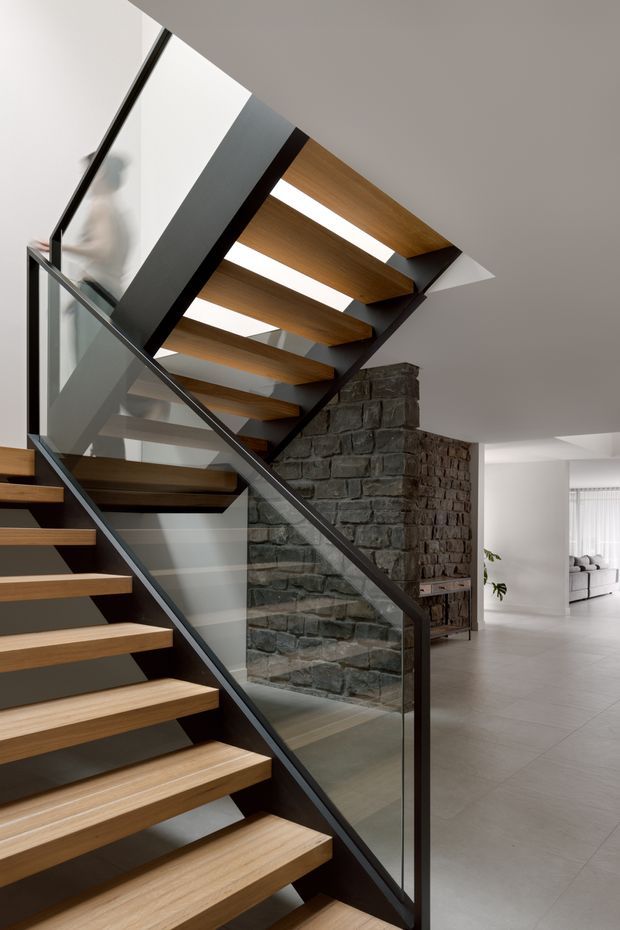 Interior view of a contemporary open-riser staircase with warm timber treads, dark metal stringers, and clear glass balustrades. A substantial exposed rough-stone masonry wall forms a feature beside the stair, with a bright open-plan living area visible beyond. The space has a restrained neutral palette, clean geometric lines, and abundant natural light.
