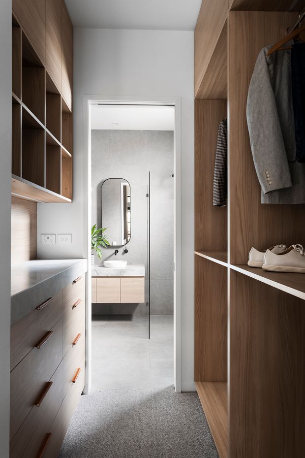 A compact, carefully fitted walk-in wardrobe or dressing area with warm timber cabinetry on both sides, open shelving, hanging rails, drawers and a light benchtop. The space opens directly into a contemporary bathroom with large grey wall and floor tiles, a frameless glass shower, floating timber vanity and rounded-edge mirror. Grey carpet covers the wardrobe floor, while white walls and ceiling keep the narrow space bright.