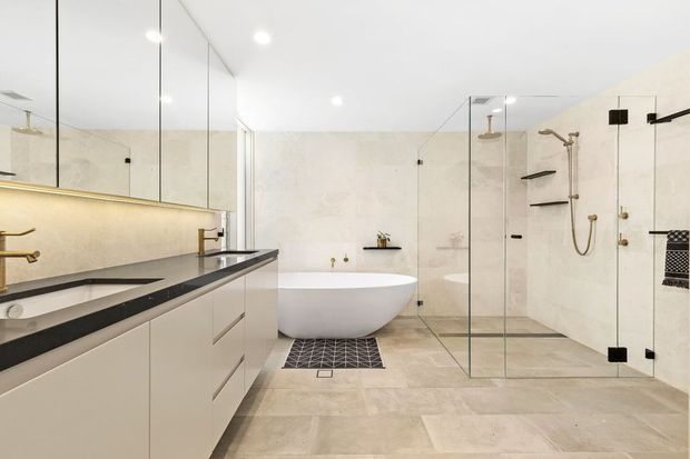 Bright, spacious contemporary bathroom with a freestanding white bathtub, double vanity, large mirrored wall, pale stone-look wall and floor finishes, and a generous frameless glass walk-in shower. Brushed brass fixtures and black accents provide contrast.