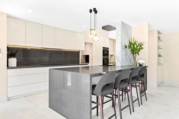 A bright, contemporary kitchen with full-height white cabinetry, a dark stone splashback, and a large grey stone island with integrated seating for five. The space features pendant lighting, built-in appliances, a dramatic veined marble-look feature wall, open shelving, and pale stone flooring.