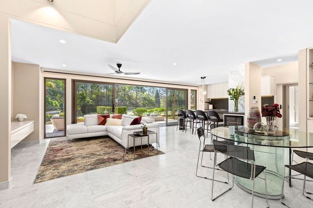 Bright, open-plan contemporary interior combining a living room, dining area and kitchen. The space features pale marble-look flooring, white walls and ceiling, extensive floor-to-ceiling glazed doors overlooking greenery, a white sectional sofa, glass dining table, dark wire dining chairs, and a compact kitchen with island seating. Recessed lighting and a ceiling fan contribute to the clean, airy presentation.
