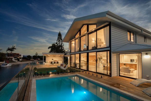 Twilight exterior view of a contemporary multi-level residence beside a swimming pool and canal or waterfront setting. The home features a dramatic glazed double-height gable end, extensive black-framed floor-to-ceiling windows, white horizontal weatherboard cladding, a low-pitched roof, and a timber pool deck. Warm interior lighting is visible through the glazing, with landscaped outdoor areas and neighbouring homes in the background.