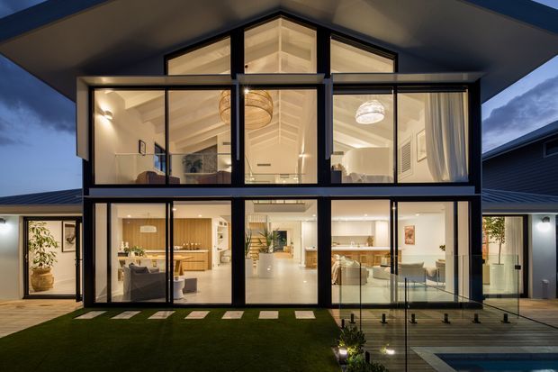 Twilight exterior view of a contemporary two-storey home with a strongly glazed central facade, dark-framed floor-to-ceiling windows, and a pitched central roof form. Warm interior lighting reveals an open-plan kitchen and living areas, sculptural pendant lights, and a bright upper-level room. A small landscaped lawn, stepping stones, timber decking, glass balustrade, and part of a pool are visible in the foreground.