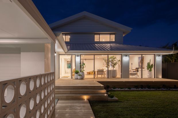 Twilight exterior view of a contemporary two-storey home with a pitched metal roof, horizontal weatherboard cladding, extensive illuminated glazing, and a raised timber deck connected by timber steps and walkway. A decorative geometric breeze-block screen forms the foreground boundary, with warm interior lighting and visible living spaces beyond the glass.
