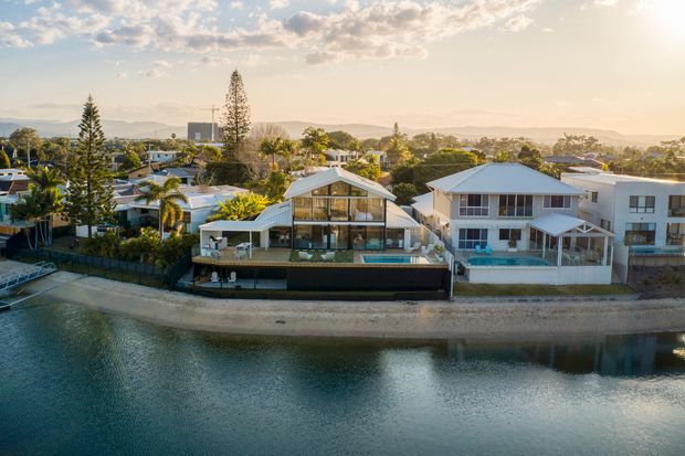 Aerial exterior view of a group of waterfront suburban homes along a calm canal or river. The central multi-level house has a prominent gabled roof, broad glazed upper facade, balcony or terrace, and a dark lower level beside the water. Adjacent light-coloured homes feature pitched roofs, balconies, and swimming pools. Warm late-afternoon light, distant hills, mature vegetation, and reflective water create a relaxed residential setting.