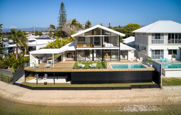 Aerial exterior view of a contemporary multi-level waterfront residence with a prominent gable roof, extensive glazing, black lower-level cladding, timber deck, covered carport, landscaped outdoor areas, and a swimming pool along the front edge of the property. Neighboring homes and a calm waterway are visible beyond the site.
