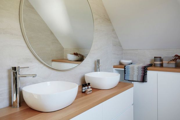 Contemporary bathroom vanity with two white vessel basins, a timber benchtop, white built-in cabinetry, chrome tapware, and a large round mirror. Pale textured stone-look wall surfaces and soft neutral styling create a clean, calm appearance.
