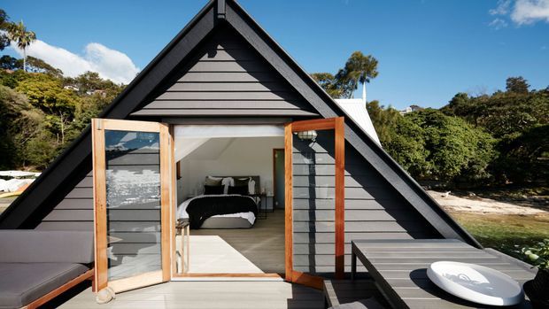 Exterior view of a compact A-frame cabin with a steep dark gabled roof and grey horizontal cladding. Large timber-framed glazed doors open onto a small deck, revealing a simple bedroom inside. The cabin is surrounded by dense vegetation and appears positioned beside a rocky waterfront.