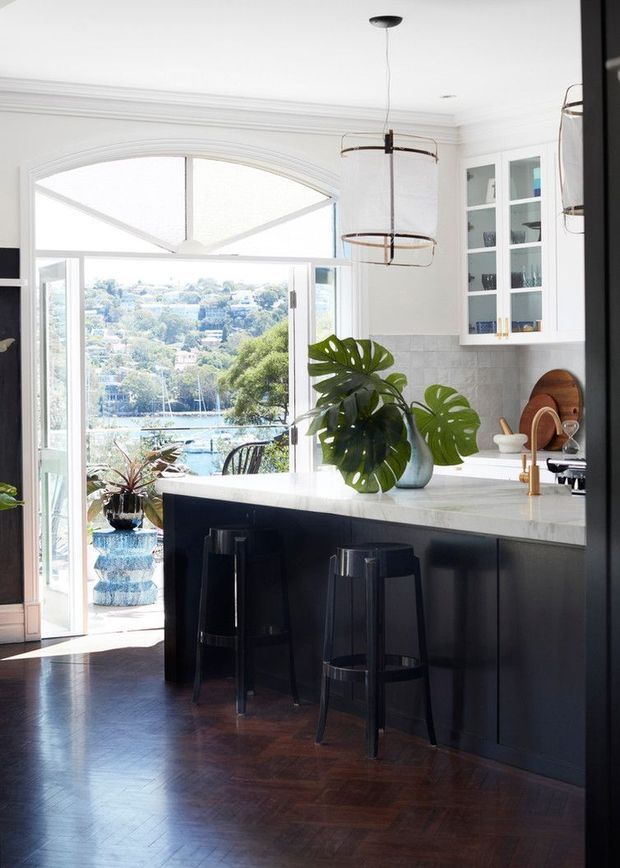 Bright, refined kitchen interior with a large marble or light stone island, dark cabinetry, black bar stools, white glass-front upper cabinets, and brass-toned tapware. A glazed door opens to a balcony or outdoor area with a leafy waterfront or hillside view. The room features dark herringbone timber flooring, white walls with decorative cornices, tiled splashback, pendant lighting, and abundant tropical foliage.
