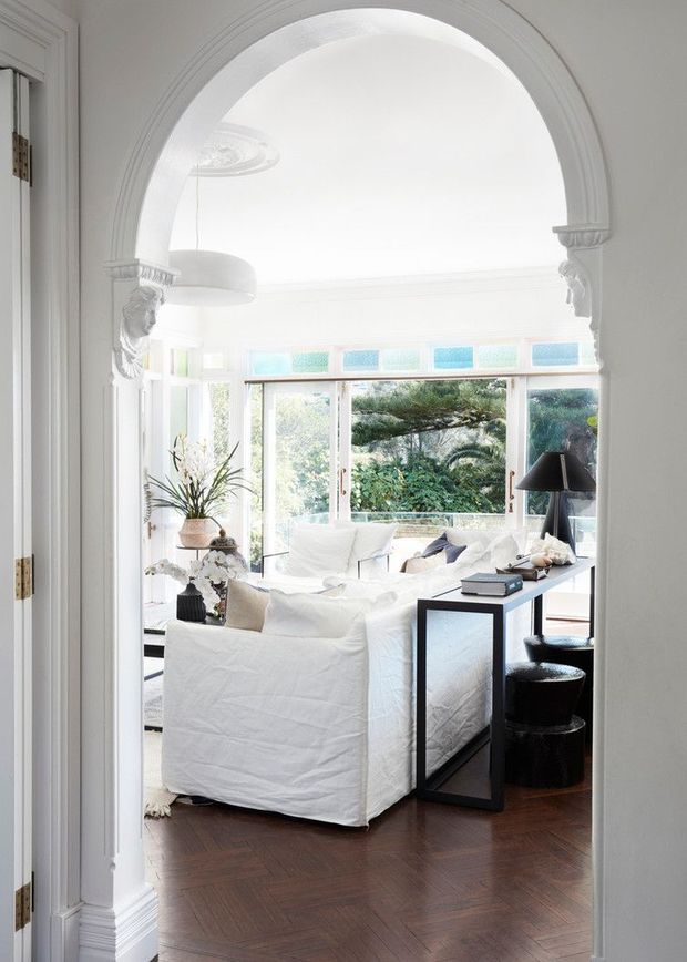 Bright, elegant living room viewed through a wide ornate arched opening. The room features white slipcovered sofas, dark timber flooring, a slim black console table, black lamps and stools, decorative wall mouldings, and large windows with coloured transom glass overlooking greenery. The overall atmosphere is airy and refined, combining classical architectural detailing with a clean contemporary interior.