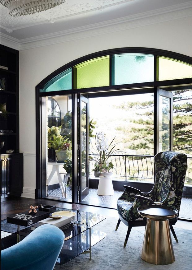Elegant living room with a dramatic arched black-framed glazed doorway opening onto a balcony or terrace. The room features ornate decorative ceiling mouldings, dark built-in shelving, a blue upholstered sofa, patterned floral armchair, glass coffee table, and a brass drum side table. Coloured translucent clerestory glazing adds a refined period character, while abundant plants and bright outdoor views create an airy indoor-outdoor connection.