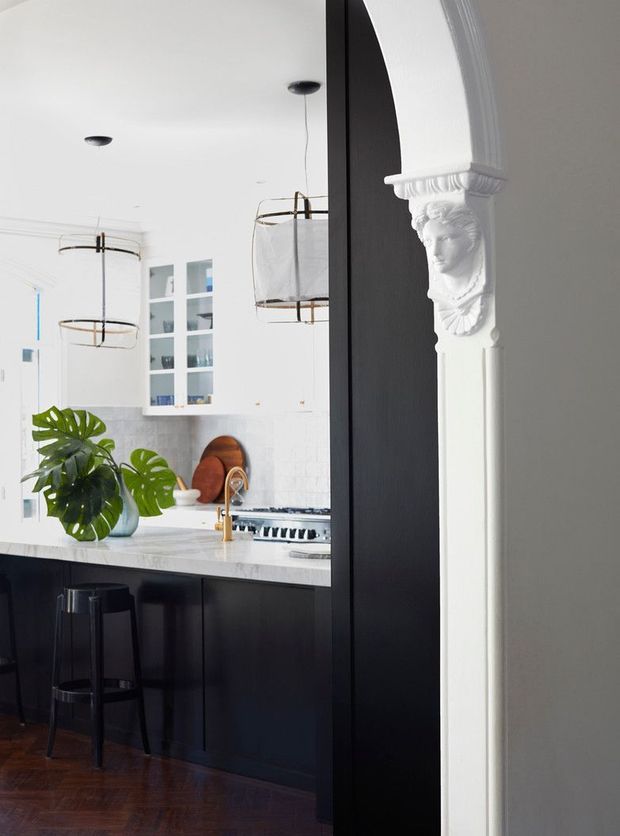 A bright, refined kitchen viewed through a wide opening, featuring matte black cabinetry and island, a pale stone benchtop, white cabinetry and tiled walls, brass tapware, black counter stools, and elegant white decorative architrave with a classical bust detail. Pendant lights and a leafy plant add visual interest, while dark timber parquet flooring anchors the space.