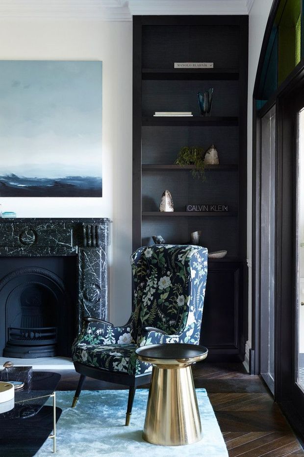 A refined living room corner featuring a dark marble fireplace, built-in black shelving, a floral upholstered wingback chair, a small metallic pedestal table, and a large abstract landscape artwork. Dark timber floors, black joinery, and generous glazing create a moody yet elegant interior.