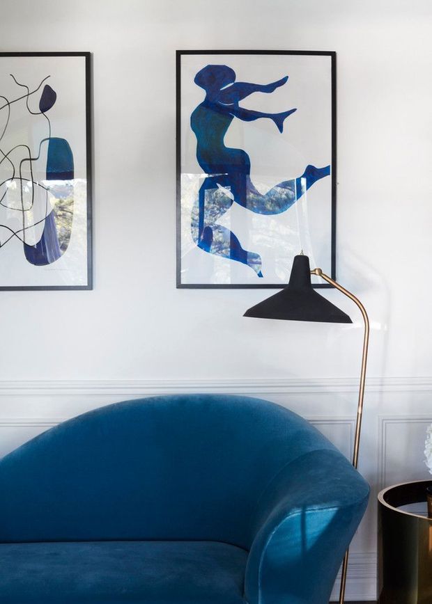 A stylish living room vignette with a deep blue curved sofa, white walls with decorative wainscoting, framed abstract blue artwork, and a sculptural black floor lamp with a slim brass stem. A small dark side table is partially visible at right. The room has a refined, high-contrast palette and abundant natural light.
