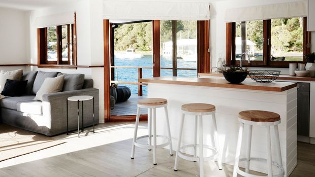 Bright open-plan interior featuring a white kitchen island with a timber benchtop and three white stools, adjoining a living area with a grey sectional sofa. Large timber-framed windows and glazed doors provide views toward a swimming pool and greenery, while white cabinetry, light flooring, and timber accents create a fresh, relaxed atmosphere.