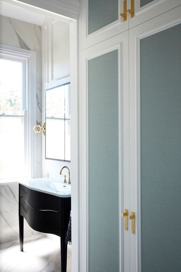 Bright, refined bathroom viewed through partially open paneled doors. The room features a black curved vanity with a white integrated basin, brass tapware and hardware, pale marble-look wall and floor surfaces, tall windows, and blue-grey textured inset wall panels framed in white cabinetry or moulding.