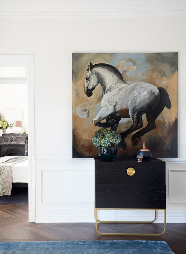 A refined interior vignette featuring a black wood console cabinet with a brass circular handle and brass-colored tubular base, topped with a potted plant and decorative vessel. The cabinet sits against a white wall with traditional raised-panel wainscoting and trim. Above it hangs a large equestrian artwork, while a partial adjoining room with a fireplace is visible through the opening on the left. Dark herringbone timber flooring and a blue rug add contrast.