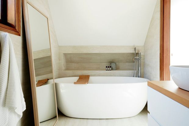 Bright contemporary bathroom featuring a freestanding white bathtub, pale timber-look wall and floor finishes, a wall-mounted bath faucet and hand shower, a large leaning mirror, and light built-in vanity cabinetry with a timber benchtop. Warm timber-framed openings and a white towel are visible at the edges.