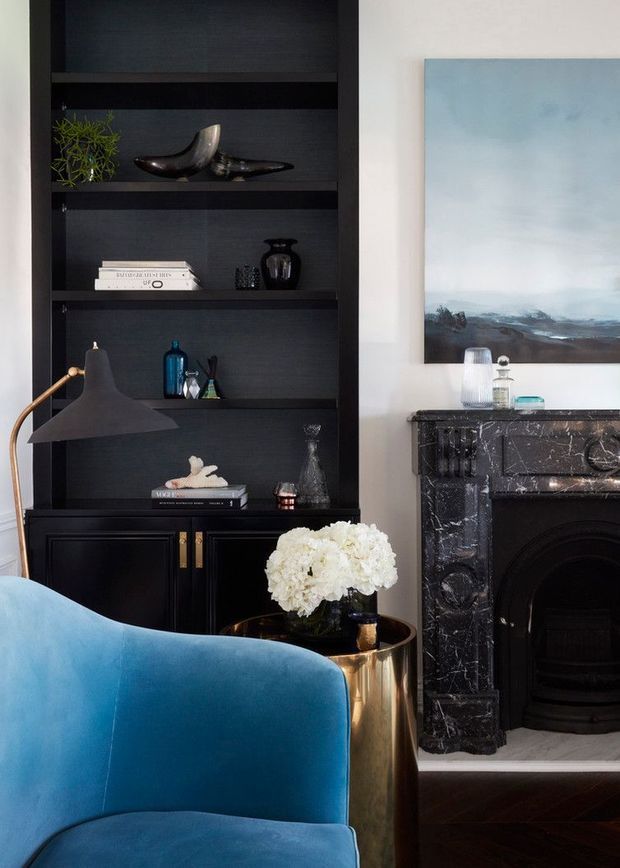 A refined living room vignette featuring a deep blue upholstered armchair in the foreground, a black built-in shelving unit displaying decorative objects and books, and a dark ornate marble fireplace with a painted landscape above it. A brass-toned side table, sculptural lamp, and white floral arrangement add contrast to the rich, sophisticated palette.