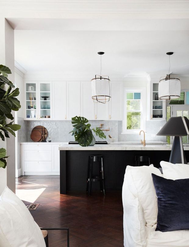 Bright, refined open-plan kitchen viewed from an adjacent living area. The kitchen features white shaker-style cabinetry with glass-front display doors, a pale patterned tile backsplash, a long black island with seating, brass-toned tapware, dark timber herringbone flooring, and two oversized pendant lights. White sofas and a dark navy cushion occupy the foreground, while leafy indoor plants add an organic accent.