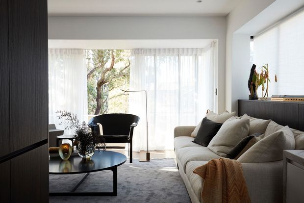 Bright contemporary living room with a large cream sectional sofa, dark accent cushions, a round black coffee table, sculptural armchair, floor lamp, sheer full-height curtains and abundant glazing overlooking trees. Dark vertical timber cabinetry and a low built-in console contrast with the light walls, carpet and upholstery.