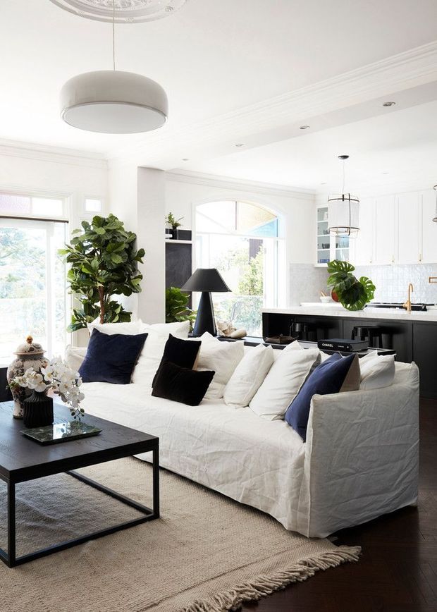 Bright open-plan living room with a large white slipcovered sectional sofa, dark coffee table, neutral rug, black accents, and abundant natural light. A contemporary black-and-white kitchen with island, pendant lighting, white cabinetry, and timber flooring is visible behind the seating area. Decorative ceiling mouldings and a ceiling medallion add traditional architectural detailing.